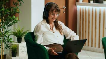 antiguo mujer acecho película en computadora portátil, escuchando a favorito televisión espectáculo en salón zona a hotel. retirado turista utilizando auriculares y computadora a pasar hora en internacional vacaciones, esperando vestíbulo. foto