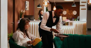 Bellhop serving coffee cup to woman client relaxing in lounge area, senior hotel guest enjoying refreshment and reading book. Employee providing all inclusive services on holiday. photo