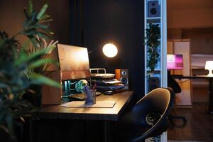 Work desk with computer and house plants in empty home studio interior with warm lighting. Apartment illuminated by neon lights with 3D rendered animations running on powerful PC monitors photo