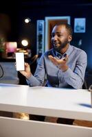 BIPOC content creator holding isolated screen smartphone in his hands, talking about specifications to online audience. Internet show host presenting mockup mobile phone features photo