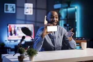 Tech engineer presents mockup phone in entry level low budget price range, filming from dimly lit home studio. Viral tech content creator reviews isolated screen smartphone in front of online audience photo