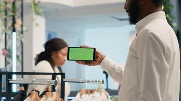 African american shopper holding green screen smartphone in cheap thrift shop with vintage clothes. BIPOC client with isolated screen chroma key mobile phone in second hand store photo