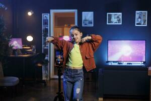 pequeño niña haciendo contenido creación para niños, utilizando teléfono inteligente en trípode a Produce danza videos para en línea transmisión plataformas linda niño creando tendencias coreografía con Teléfono móvil foto