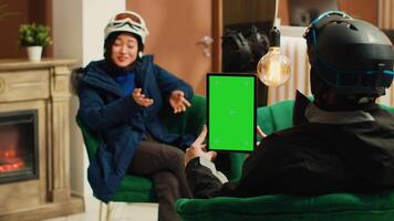 Traveller shows isolated greenscreen on tablet while she sits with skiing partner in lounge area, preparing for winter sport with gear. Man holding gadget with blank chromakey mockup template. photo