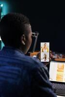 Black woman holding mobile device is on video call with caucasian clients while using her digital laptop. African american freelancer is virtually communicating with colleagues on her smartphone. photo