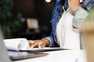 Close up of black person reviewing documents at desk while speaking on mobile device. Selective focus on african american female individual with finger on clipboard, checking and analyzing paperwork. photo