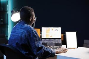 While browsing on laptop, african american woman sits at desk with tablet showing isolated white screen. Female freelancer using personal computer next to digital device displaying mockup template. photo