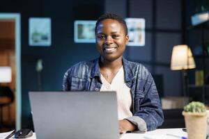 negro mujer trabajando a su ordenador portátil en un moderno departamento, estudiando en línea y investigando información para su proyecto. sonriente africano americano blogger utilizando personal computadora mientras mirando a cámara. foto