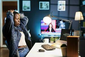 Black woman with hands on head sadly looking at laptop screen showing game over. Upset african american female gamer sitting at desk in apartment after losing video game on portable computer. photo