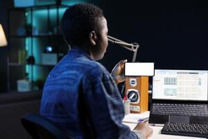 African american woman works from home on laptop, holding mobile device with isolated white screen. Female freelancer using wireless computer while grasping smartphone with blank mockup template. photo