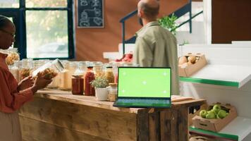 Greenscreen on device in eco store with crates of fresh homegrown products and bulk items in jars on shelves. Laptop showing isolated template with mockup chromakey display. photo