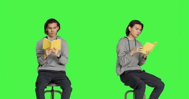 Asian person reading literature book over greenscreen backdrop, enjoying lecture while he sits on chair. Young adult reads fiction novel, being interested in old school hobbies. photo