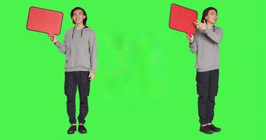 Young guy shows speech bubble in studio, presenting isolated copyspace on billboard icon against greenscreen. Asian man working on creating marketing commercial, advertisement. photo