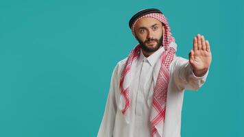 Middle eastern model doing stop symbol with palm raised, expressing negativity and rejection on camera. Young adult showcasing refusal, being displeased with something in studio. photo