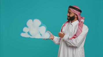 Middle eastern adult holds cloud icon on camera, showcasing blank billboard icon in natural layout. Male model in traditional muslim apparel and headdress doing new commercial advertisement. photo