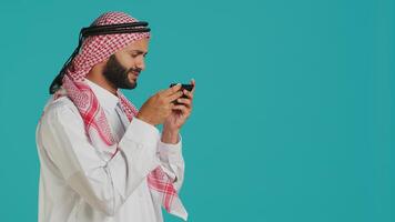 Arab man playing fun mobile videogames on phone, dressed in traditional attire with dress and headscarf, leisure activity. Middle eastern guy engaging in online gaming contest using device. photo