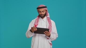 Middle eastern man checking tablet in studio, browsing websites online on digital gadget. Male person in arabic clothes using device to navigate on internet pages, texting on social media. photo