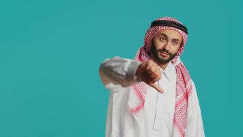 Muslim guy with ancient gown and kufiyah gives thumbs down and feels unsatisfied with stuff. Young person expresses an apparent denial and negative sign in studio, arabic model. photo