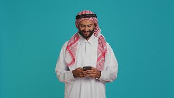 Cheerful person messaging on phone, wearing islamic religious attire and headscarf. Muslim guy dressed in traditional thobe and checkered print ghutra chatting on social media network. photo