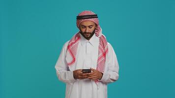 Muslim man browsing smartphone app and feeling cheerful after finding out about great news, typing texts on social media website. Middle eastern guy scrolling online pages, traditional clothes. photo