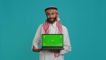 Islamic man presents greenscreen on pc, showing isolated chromakey template on laptop display. Young person holding wireless computer with mockup screen and copyspace, wearing thobe and headscarf. photo