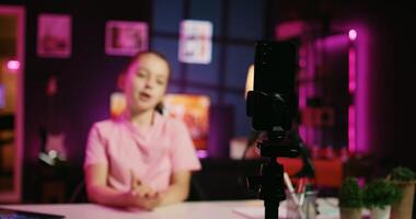 Charming small child shooting video for social media platform in living room, talking about her day. Kid entertaining followers, doing content creation in pink neon lit apartment using phone on tripod photo