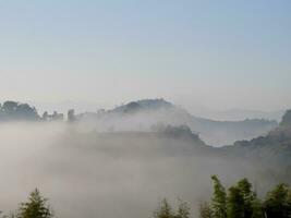 The mist flows through the mountain forest, Sun shining into tropical forest, Mist drifts through mountain ridges in the morning, slow floating fog blowing cover on the top of mountain photo