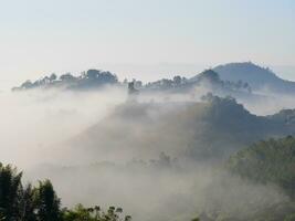 The mist flows through the mountain forest, Sun shining into tropical forest, Mist drifts through mountain ridges in the morning, slow floating fog blowing cover on the top of mountain photo