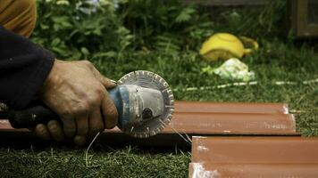 Roofer using an angle grinder machine to cut a roof tile of red color. Stock footage. Close up of male worker hands cutting a roof tile with a professional saw. photo