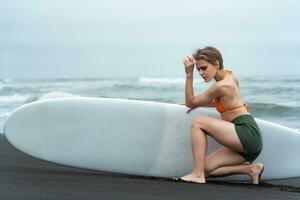 Female surfer sits in front of surfboard on beach, looking down in beautiful sexy pose photo