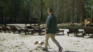 Man walks with dog on beach in woods. Stock footage. Man walks with his dog on sand at resort in summer. Man with dog walking on shore of pond in forest on sunny summer day photo