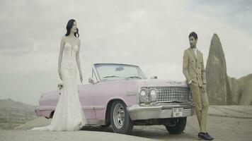 hermosa al aire libre Disparo siguiente a un rosado coche. acción. un elegante Pareja con un morena niña en un blanco vestir y un hombre en un traje son caminando en frente de un rosado coche. foto