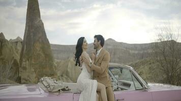 Beautiful bride and groom in wedding dress and suit near pink vintage cabriolet car. Action. Wedding couple standing near a convertible on a natural background. photo