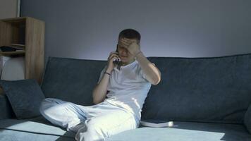 Young man is talking on phone while sitting on couch. Media. Young attractive man is talking on phone at home. Man is talking on phone call and telling something photo