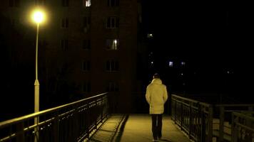 peatonal puente con un solitario calle lámpara y un hombre cruce eso a noche. valores imágenes. posterior ver de un joven hombre en blanco chaqueta cruce el puente en el ciudad a noche. foto