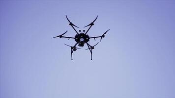 Quadcopter drone hovering in a clear blue sky, video and photo shooting concept. Clip. Black small quadrotor with digital camera and rotating blades, bottom view.