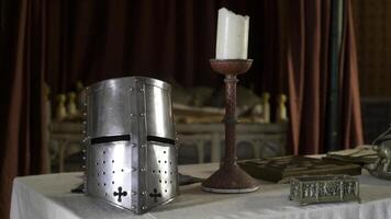 Medieval interior with details and objects. Action. Silver knight's helmet and candle stand on table in medieval interior. Objects of middle ages are on table on background of red fabric in Museum photo