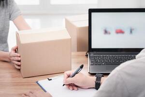 Post man use a pen write on file paper document and check online system to send parcels and women hold paper box laptop on table delivery concept work photo