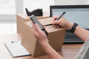 Close up hand a man use pen write on parcel box about the parcel delivery address use smartphone and laptop to register in the online system photo