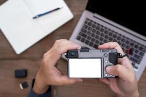 un hombre sostener cámara en mano con blanco pantalla monitor en cámara y fotografía equipo en mesa foto