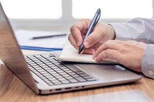 Business man write on notebook paper and use laptop computer working and analyzing with document data document for new project of business strategy at workplace. photo