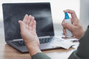 cerca arriba mano un hombre utilizar alcohol botella rociar limpiar mano y ordenador portátil computadora en el mesa primario enfermedad prevención en hogar concepto foto