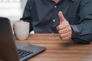 Close up hand a man thumbs up and drink coffee for work on the project successfully using computer and writing on a paper on desk in office photo