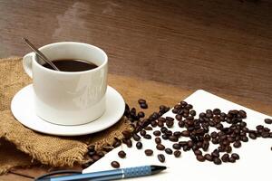 Hot Coffee in white cup and coffee beans on the note book with pen,vintage wooden table on relax concept photo