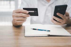 un hombre sostener crédito tarjeta y un teléfono inteligente en mano mirando inteligente teléfono con compras en línea y lápiz cuaderno papel en madera mesa foto