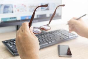A man taking off glasses, feeling eye strain after long work with desktop computer and use a pen in the hand and smartphone on the wood table photo