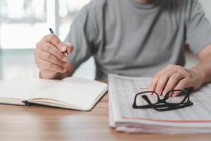 Male with a pencil writes job application information from the print book and glasses newspaper on the wood table photo