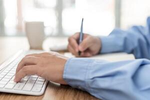 mano un hombre mecanografía en computadora escritorio teclado y utilizar un bolígrafo escribir en Nota libro en trabajando foto