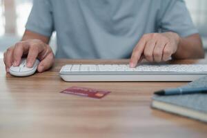 un hombre utilizar ratón escritorio computadora y teclado con crédito tarjeta bolígrafo y Nota libro en mesa trabajando a hogar concepto foto