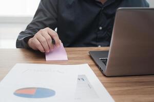 Business man write notes on paper   and use laptop computer calculate and analyzing with document data graph for new project of business strategy at workplace. photo